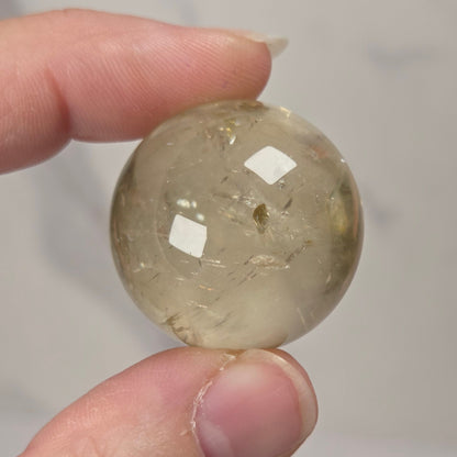 A hand holding a small, polished, translucent oval stone with natural inclusions, set against a soft, light gray marble background.
