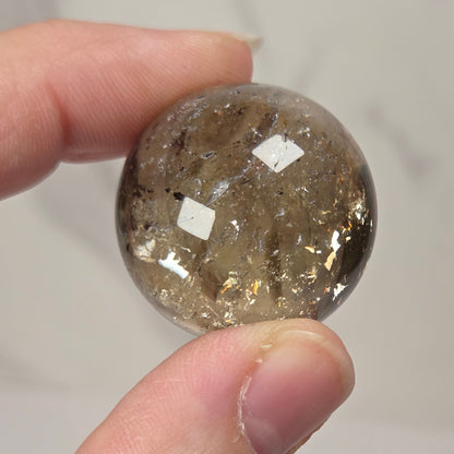 A hand holding a small, round, transparent smoky brown gemstone or crystal with visible internal inclusions against a light gray background.