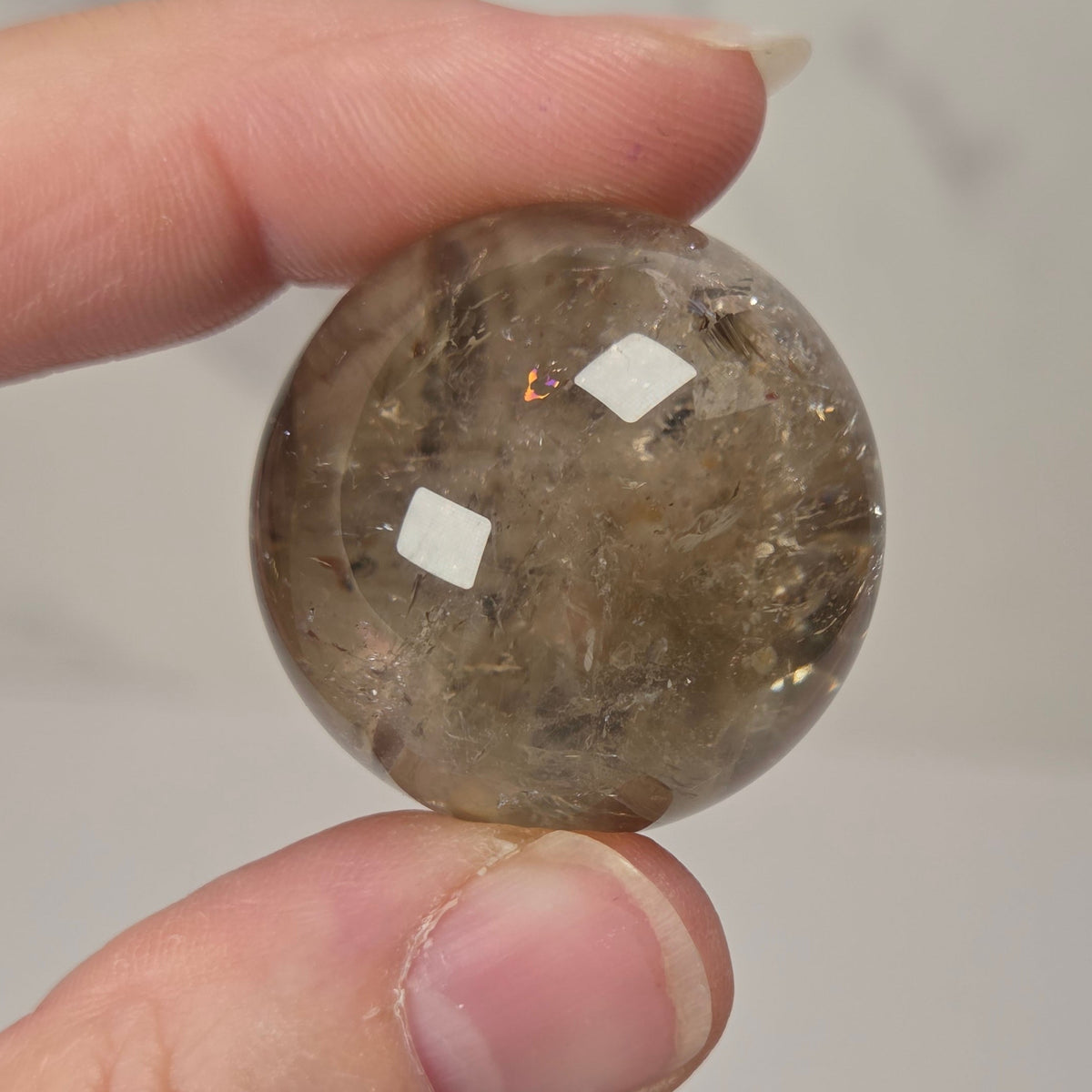 A hand holding a small, polished round gemstone with visible inclusions, set against a light gray marble-patterned background.