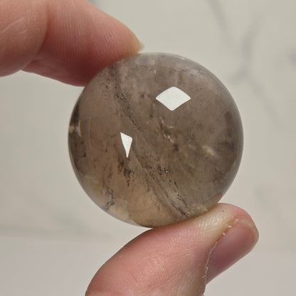 A hand holding a polished round stone bead with natural brown and gray marbling, set against a light gray marble-patterned background.