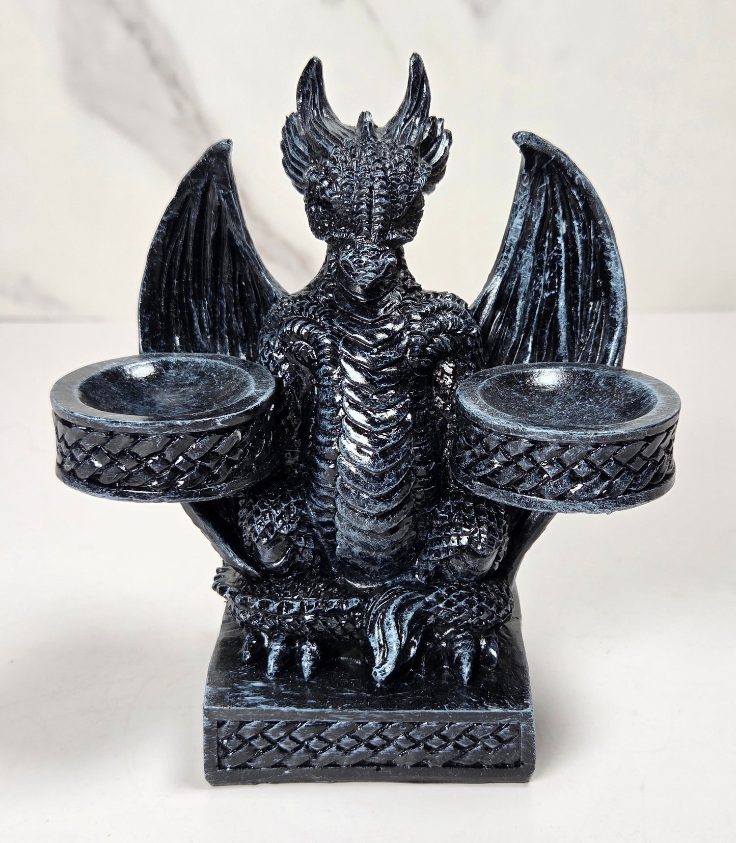 A black dragon figurine candle holder with outstretched wings and two cup-like holders on the sides, shown against a light marble-patterned background.