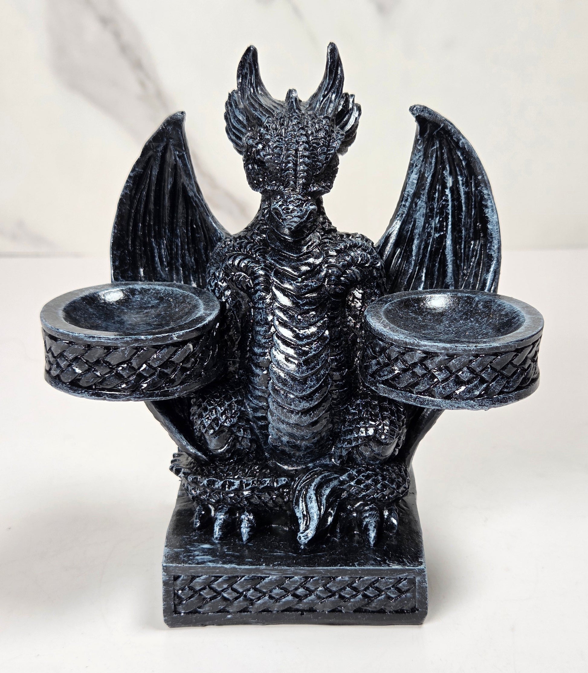 A black dragon figurine candle holder with outstretched wings and two cup-like holders on the sides, shown against a light marble-patterned background.