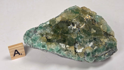 A green and yellowish crystal mineral specimen with a rough natural texture, displayed on a light off-white background next to a small wooden letter block showing the letter 'A'.