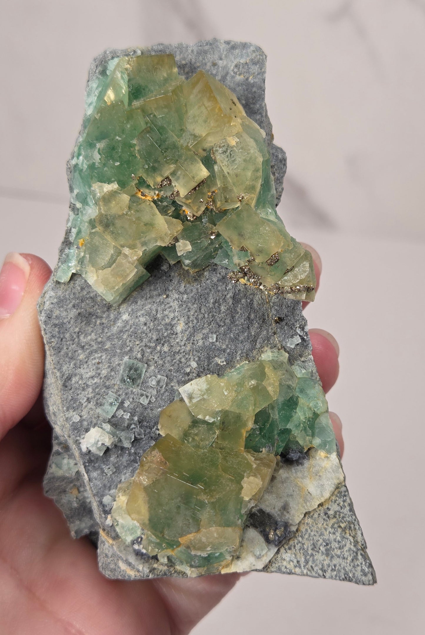 A hand holding a raw mineral specimen featuring green fluorite crystals on a gray matrix, photographed against a light marble-patterned background.