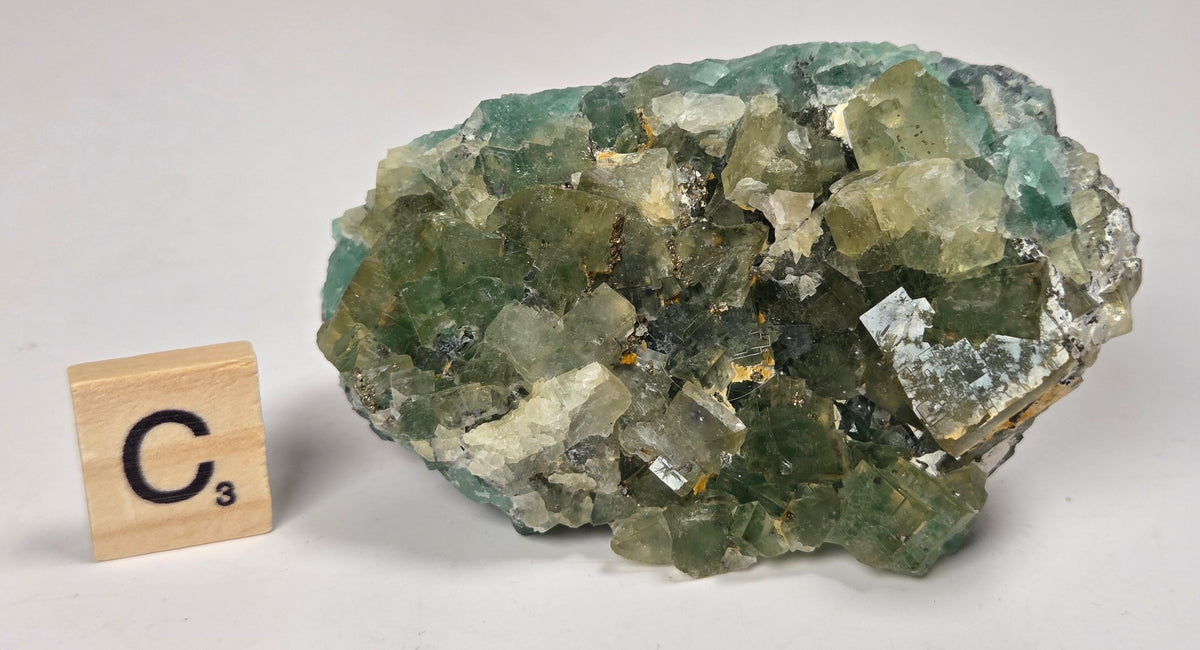 A natural fluorite crystal cluster with green, blue, and clear faceted stones, shown next to a small wooden Scrabble tile marked C3 on a light gray surface.