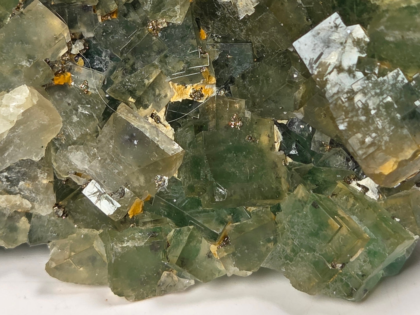 A close-up of a natural fluorite crystal cluster with green, gray, and translucent facets, showing small golden inclusions.