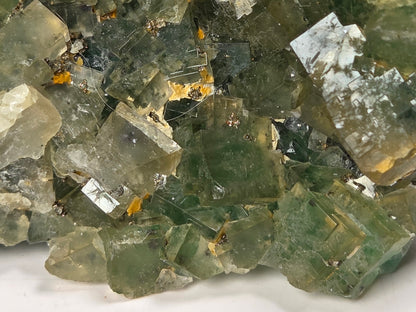 A close-up of a natural fluorite crystal cluster with green, gray, and translucent facets, showing small golden inclusions.