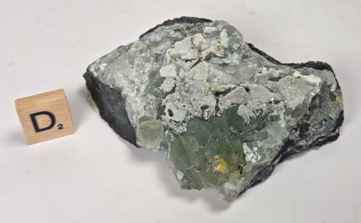 A raw mineral specimen with a green and white crystalline structure on a dark matrix, shown next to a small wooden letter tile displaying 'D2' on a plain off-white background.