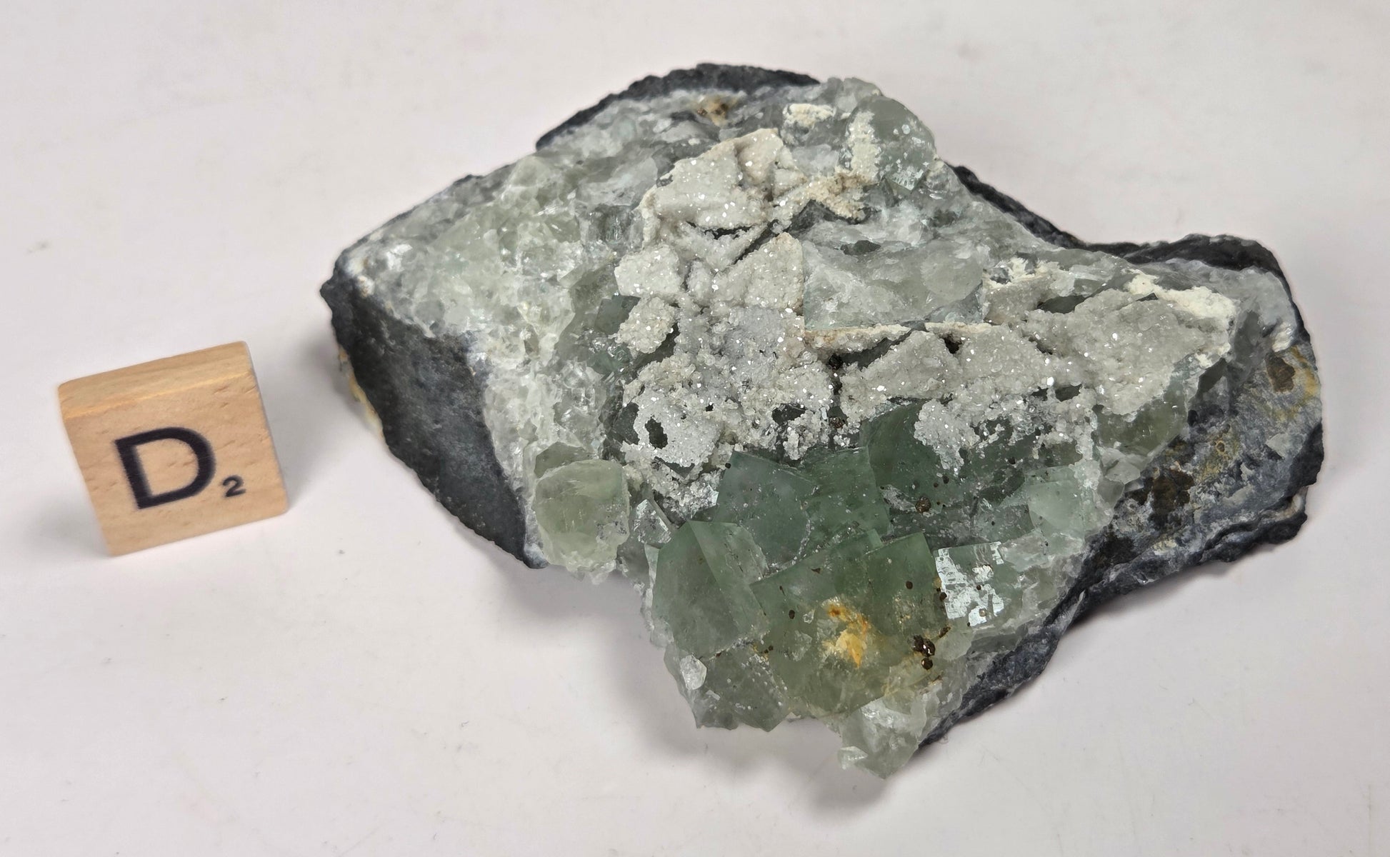 A raw mineral specimen with a green and white crystalline structure on a dark matrix, shown next to a small wooden letter tile displaying 'D2' on a plain off-white background.