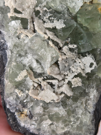 A close-up of a mineral specimen showing pale green translucent crystals over a white crystalline matrix, mounted on a dark rock base with a finger holding it at the lower left edge.
