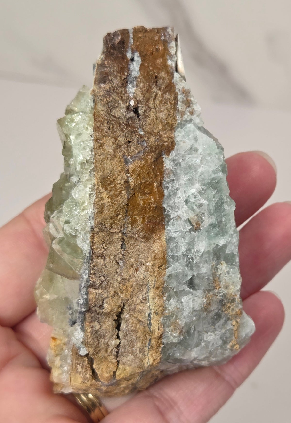 A hand holding a raw mineral specimen featuring a vertical band of brown pyrite over a pale green and white quartz matrix.