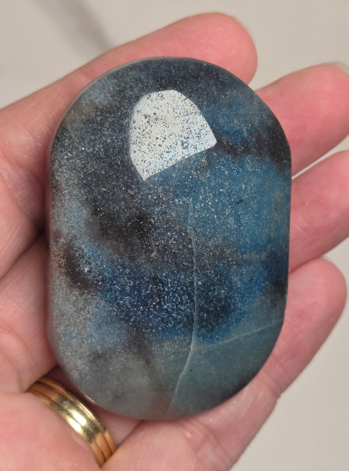 A hand holding a polished oval stone with a mottled blue, gray, and black speckled pattern, with a gold ring on the finger, against a neutral beige background.