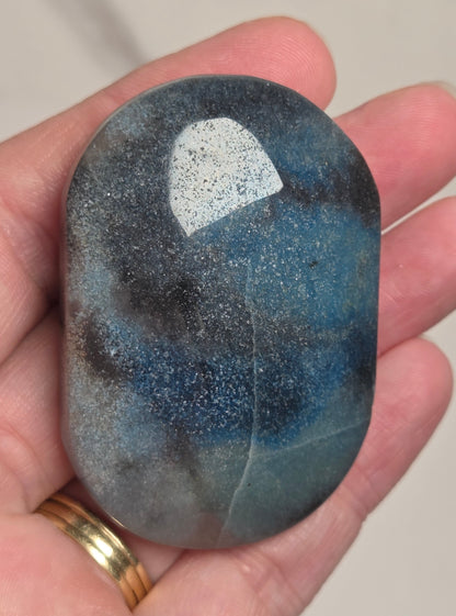 A hand holding a polished oval stone with a mottled blue, gray, and black speckled pattern, with a gold ring on the finger, against a neutral beige background.