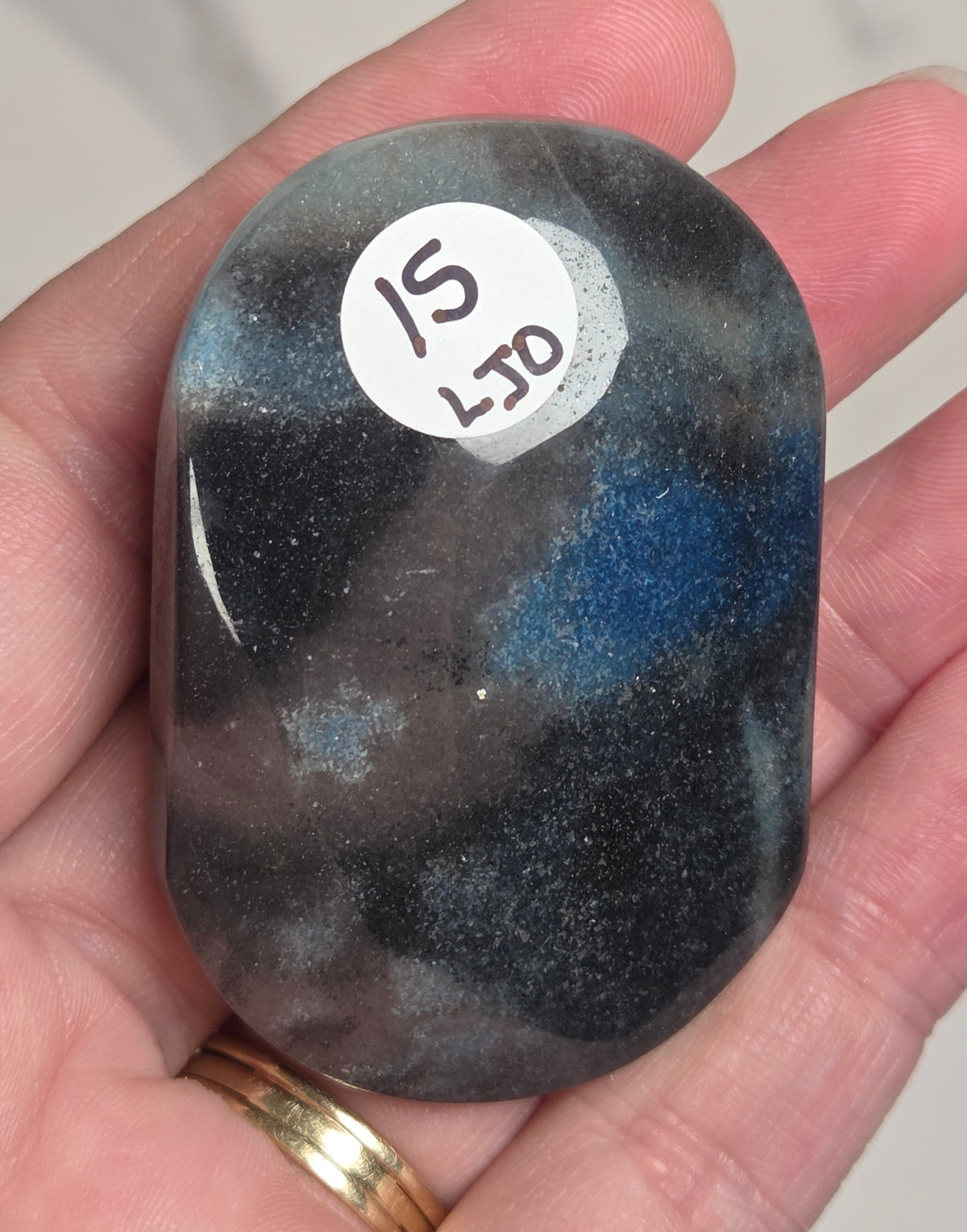 A polished tumbled gemstone with a marbled pattern of black, gray, and vibrant blue, held in a person's hand. A white circular sticker with the numbers '15 L19' is adhered to the stone's surface.