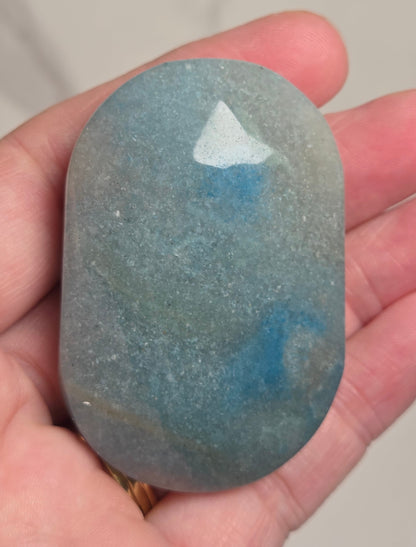 A hand holding a polished, rounded blue stone with natural marbled patterns of teal, blue, and gray.