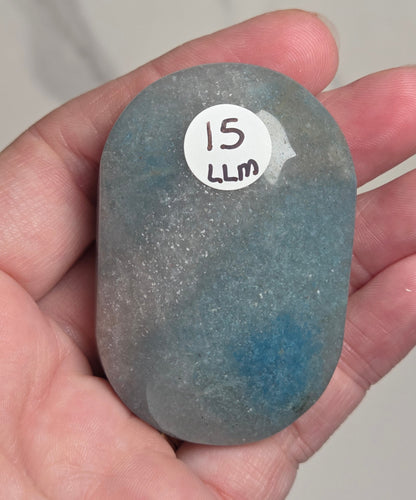 A hand holding a smooth, polished oval-shaped stone with a blue-gray color and a white sticker that reads '15 LLM'.