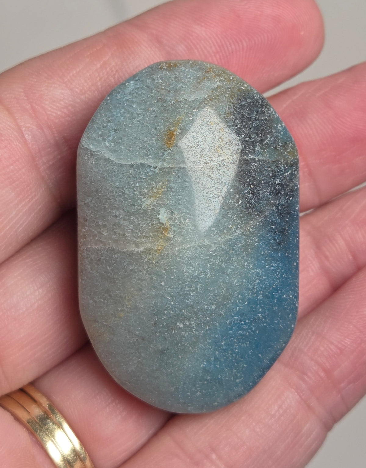 A polished oval labradorite stone held in a person's hand, showing a natural blue-gray color with a white diamond-shaped flash and subtle golden inclusions.