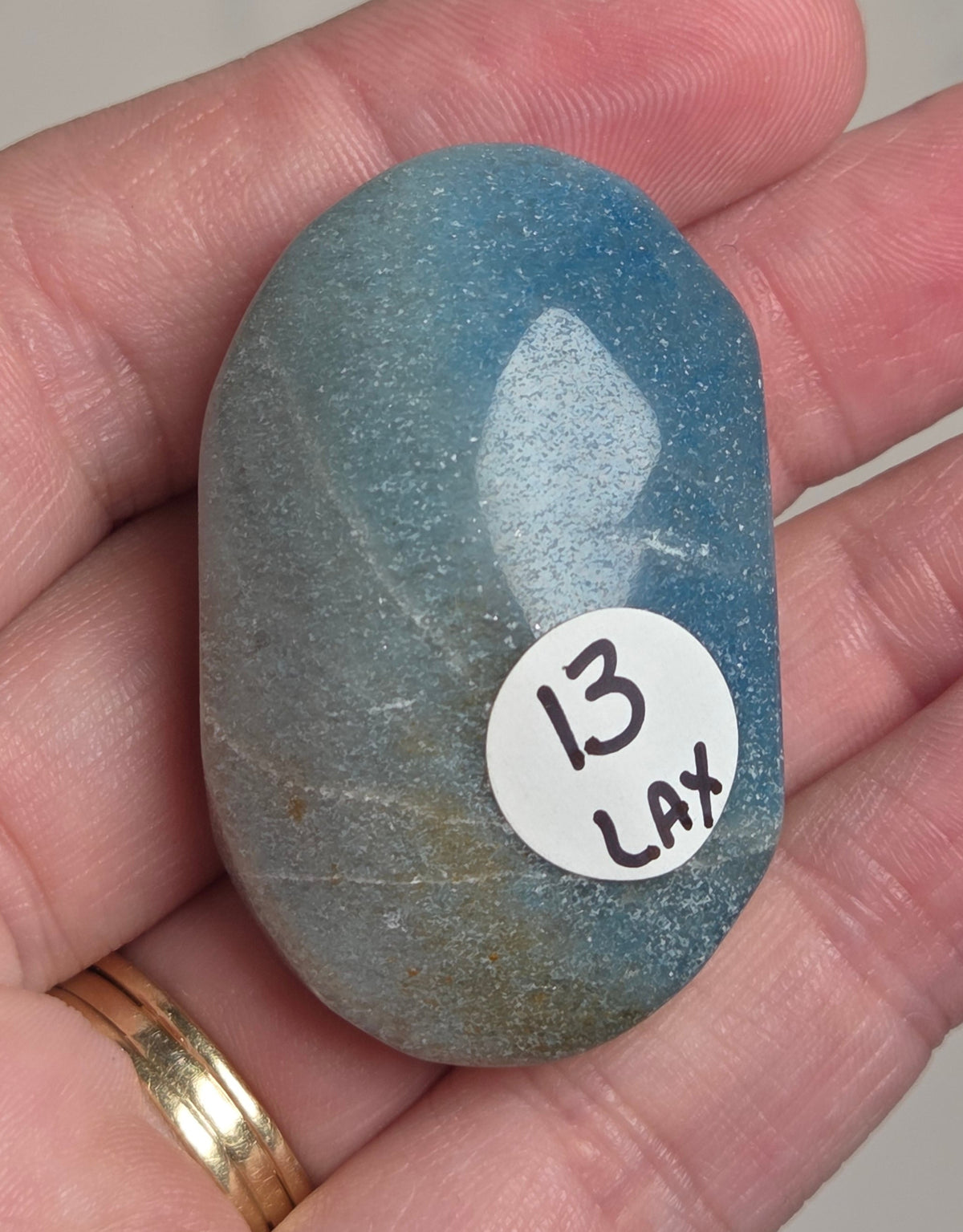 A hand holding a polished blue-gray stone with a white circular sticker that reads '13 LAX'.