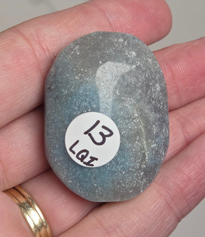A hand holding a polished, tumbled blue-gray stone with a white circular sticker that reads '13 LAZ'.