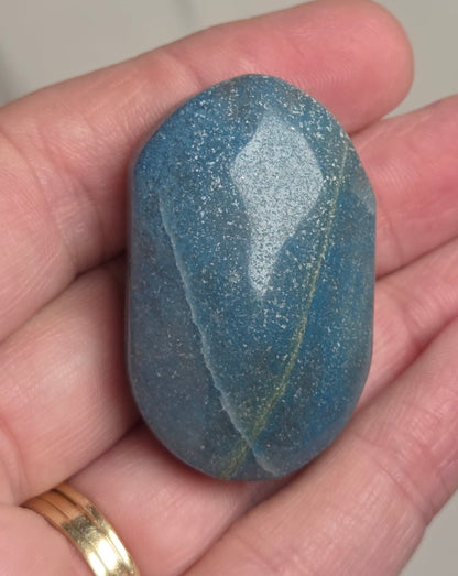 A polished blue stone with fine glitter, held in a person's hand. The stone has a rounded, egg-like shape with smooth, reflective surfaces and a subtle golden-green vein running through it.