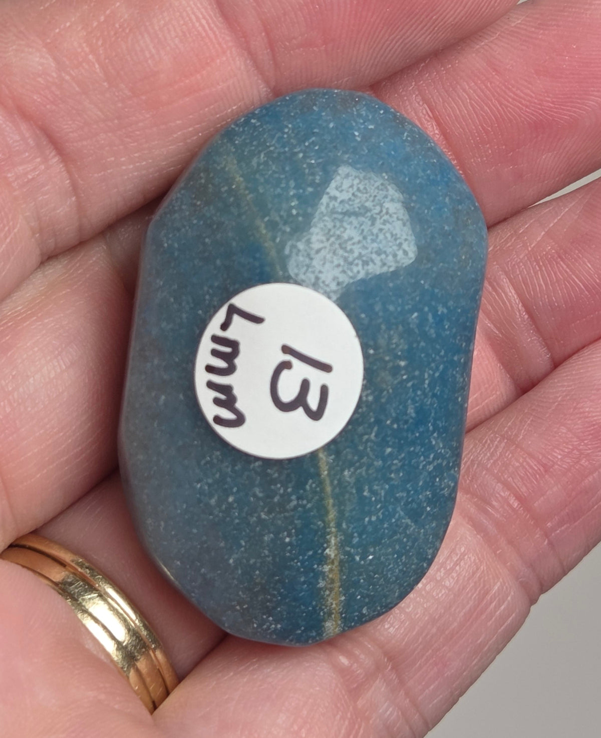 A hand holding a smooth, polished blue stone with a small white circular sticker that reads '13 Lmm'.