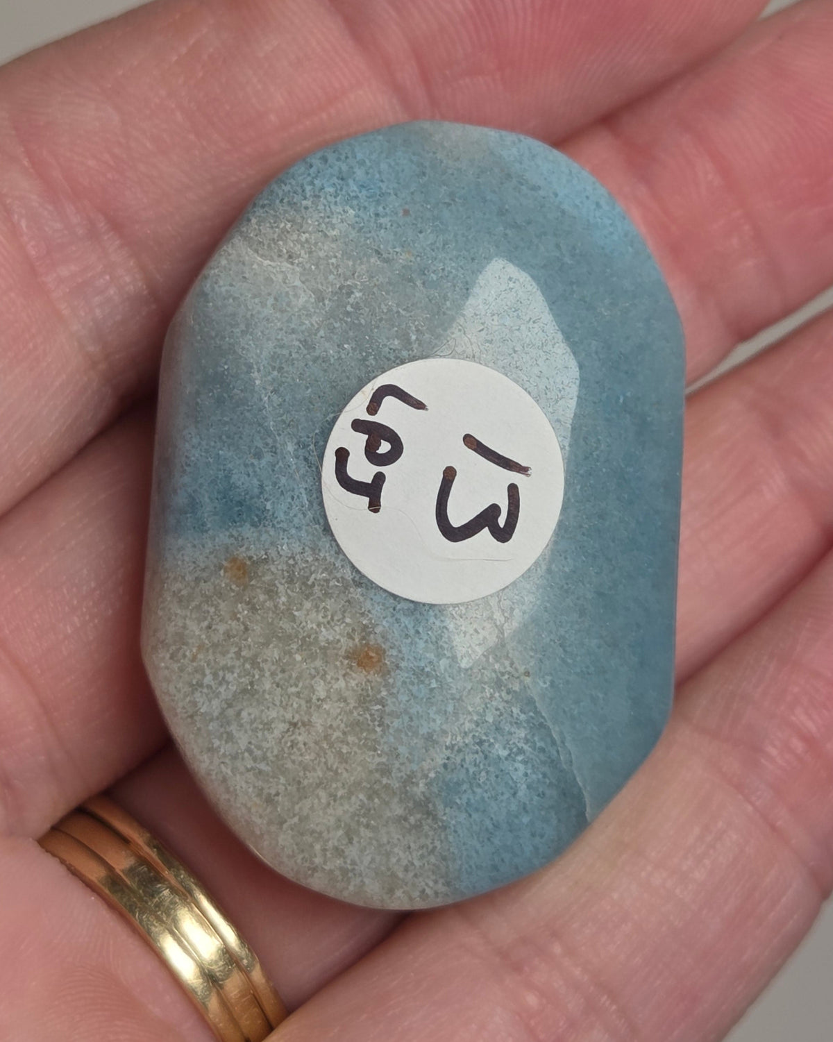A hand holding a smooth, polished blue stone with a small white circular sticker that has black handwritten characters. The hand is resting on a neutral gray surface.