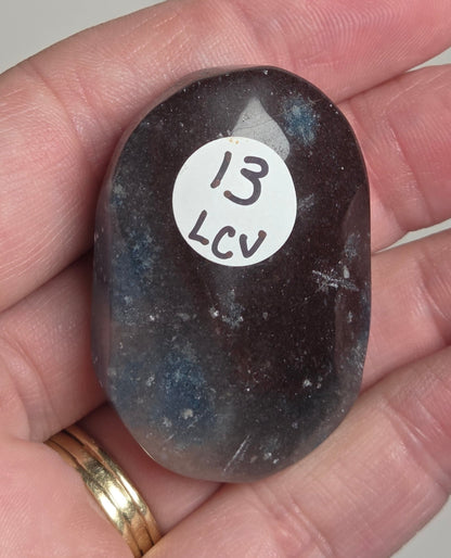 A polished dark gray to black tumbled stone with a white circular sticker reading '13 LCV' is held in a person's hand. The stone has subtle blue and white inclusions.