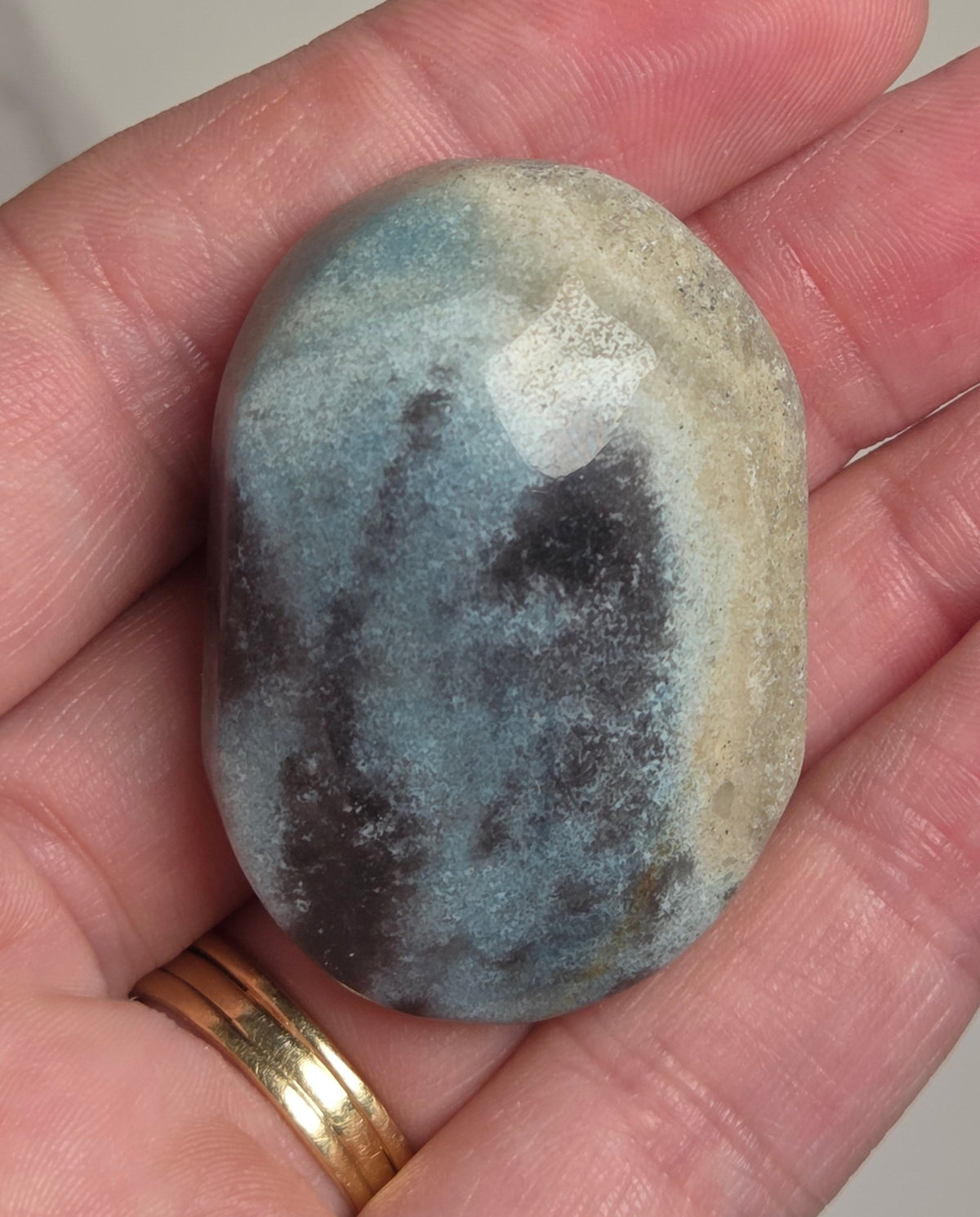 A polished oval stone with a mottled blue-gray and beige pattern, held in a person's hand. The hand has a gold ring on one finger, and the background is a soft-focus light gray marble surface.