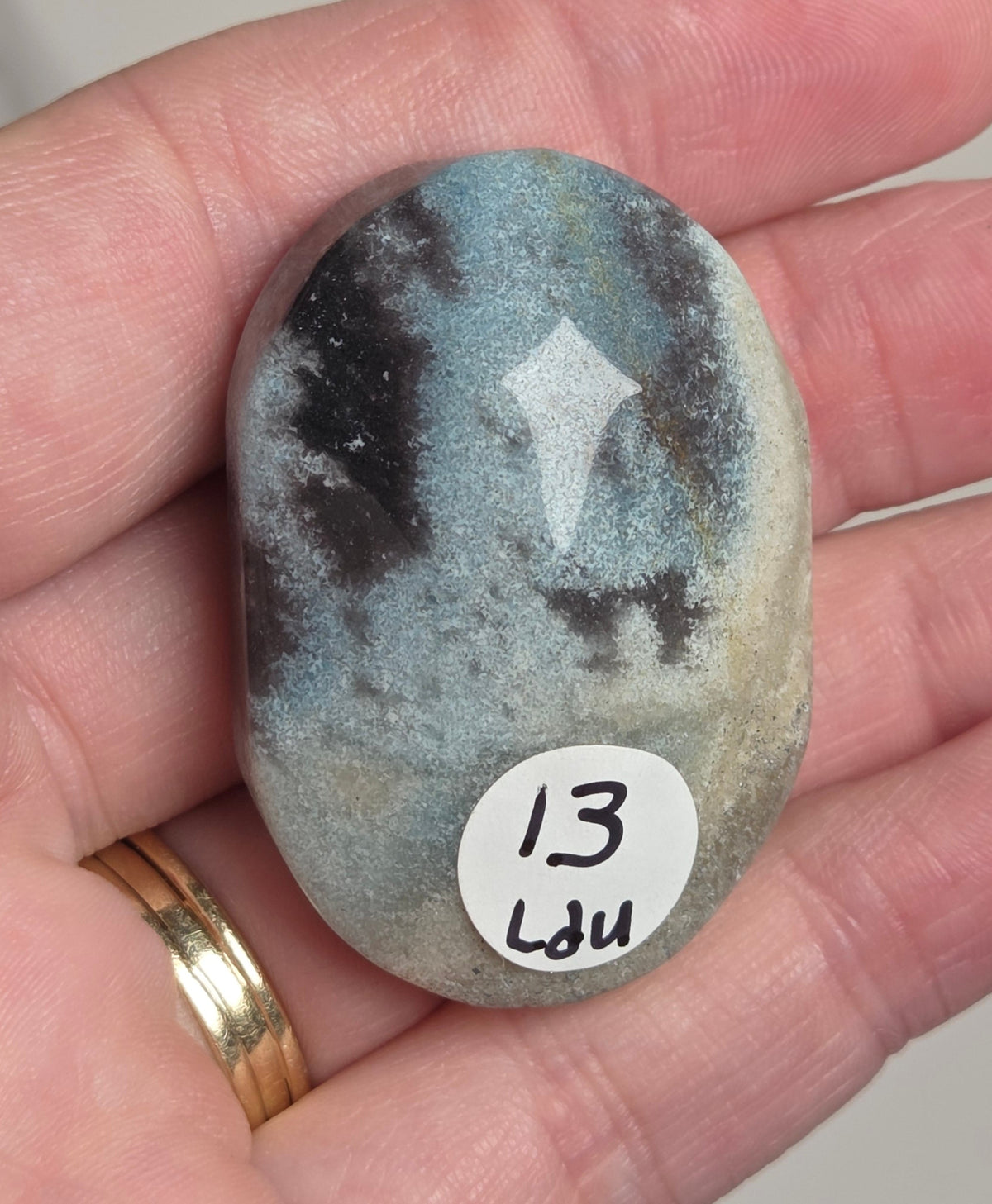 A hand holds a polished oval stone with a mottled gray, black, and blue-gray pattern and a white diamond-like inlay near the top. A small white sticker on the stone reads '13 Lau'.