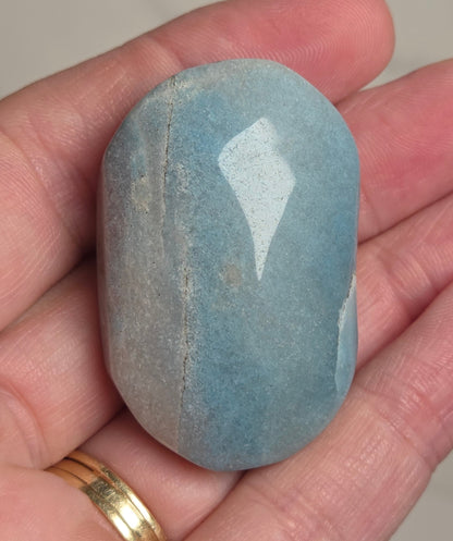 A polished light blue stone held in a person's hand, with a gold ring visible on the ring finger against a neutral light background.