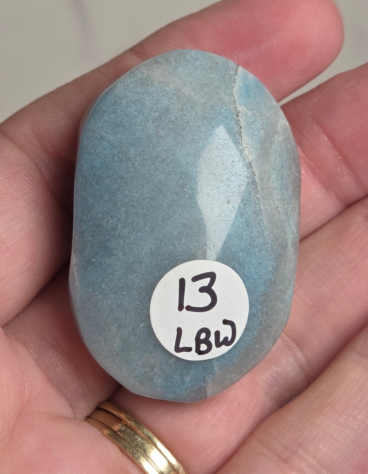 A hand holding a smooth, polished light blue stone with a white circular sticker that reads '13 LBW'.