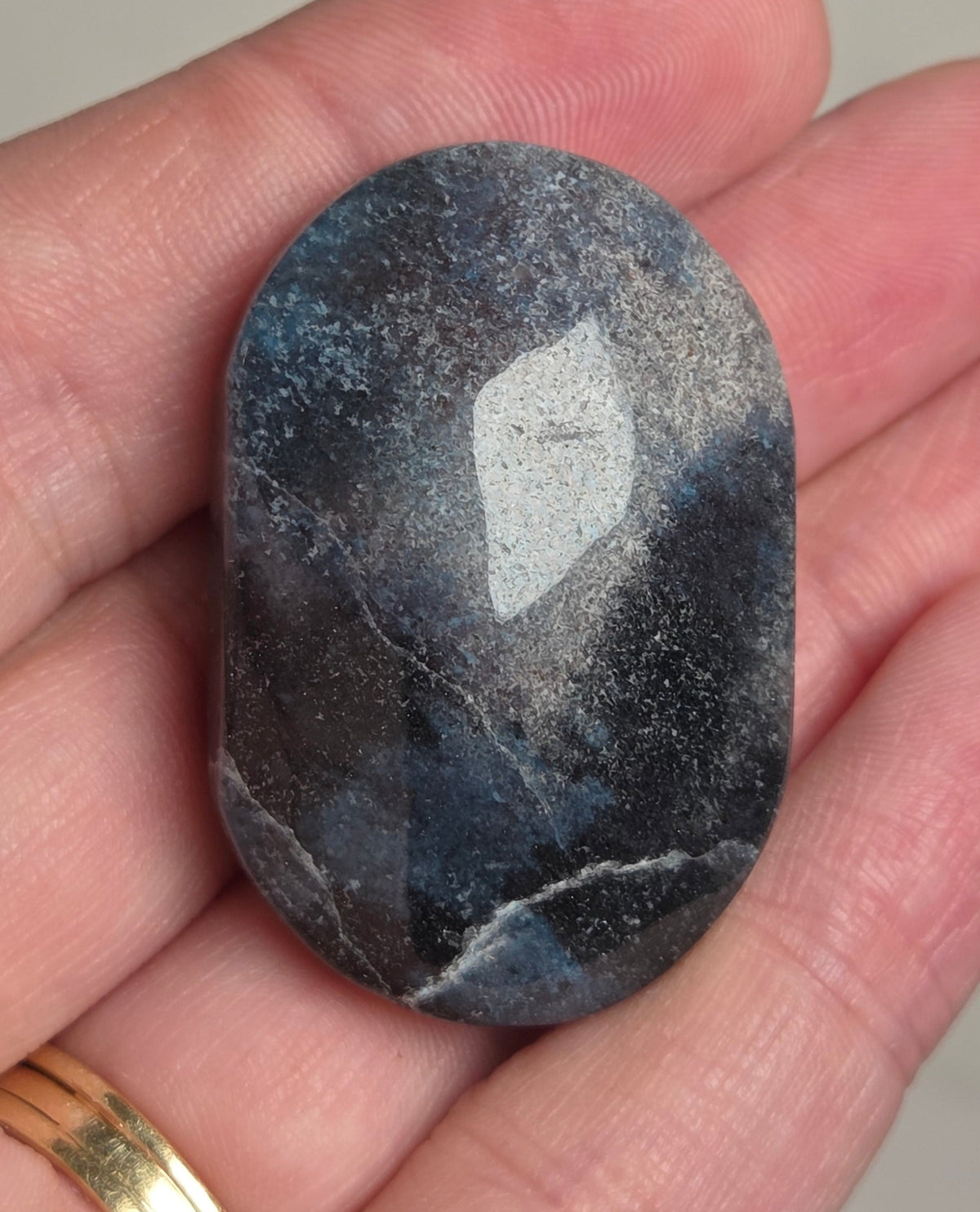 A polished oval labradorite stone with a blue-gray sheen and white flash, held in a person's hand wearing a gold ring.