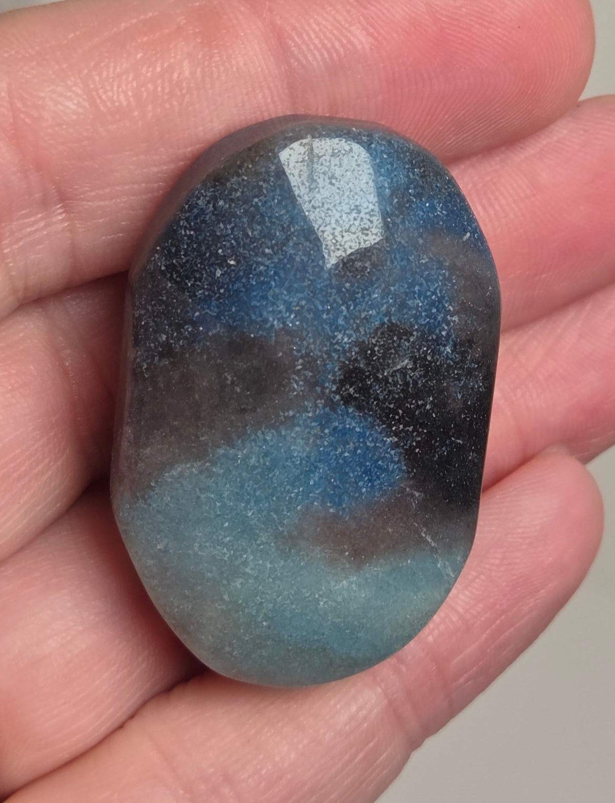 A hand holding a polished tumbled stone with a mottled blue, gray, and black marbled pattern, with a gold ring visible on the left hand.