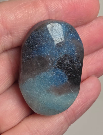 A hand holding a polished tumbled stone with a mottled blue, gray, and black marbled pattern, with a gold ring visible on the left hand.