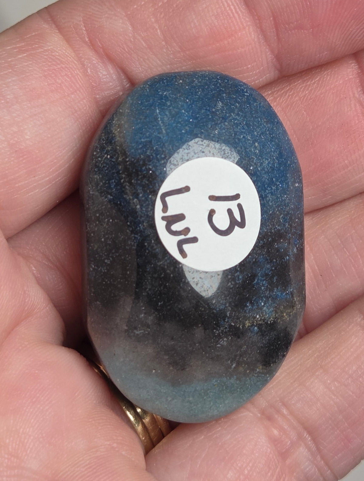 A hand holding a polished blue and black stone with a white circular sticker labeled '13 Jnd' on it.