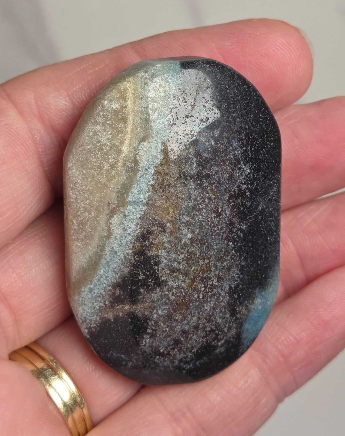A hand holding a polished, smooth stone with a marbled pattern of black, gray, beige, and light blue, wearing a gold ring on one finger against a light marble background.