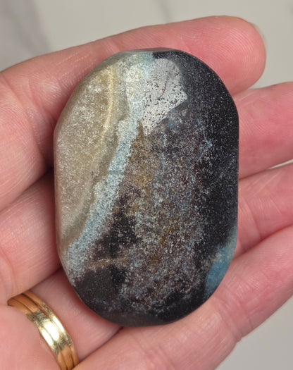 A hand holding a polished, smooth stone with a marbled pattern of black, gray, beige, and light blue, wearing a gold ring on one finger against a light marble background.