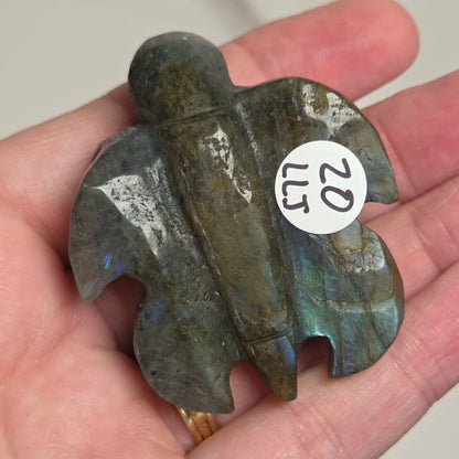 A hand holds a carved Labrador teardrop pendant with a white sticker labeled '20 LLS' on it. The pendant is made of polished labradorite showing gray, green, and blue iridescent colors.