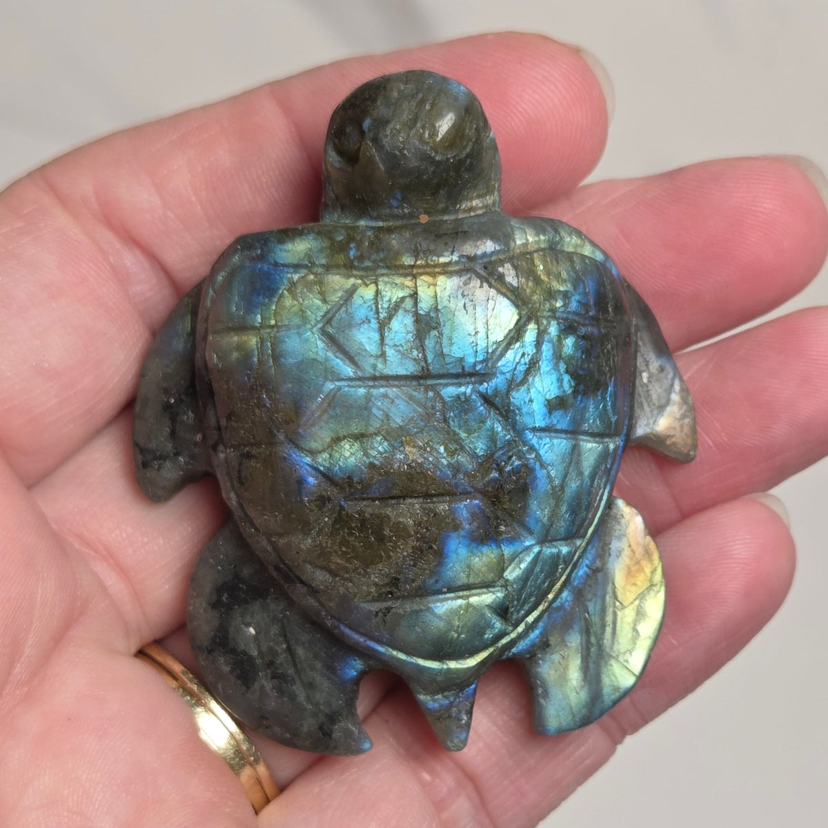A hand holding a carved sea turtle figurine made of labradorite, showing its characteristic blue, green, and gold iridescent shimmer on a light gray marble background.