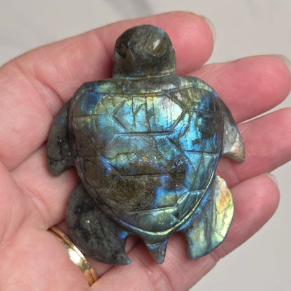 A hand holding a carved sea turtle figurine made of labradorite, showing its characteristic blue, green, and gold iridescent shimmer on a light gray marble background.
