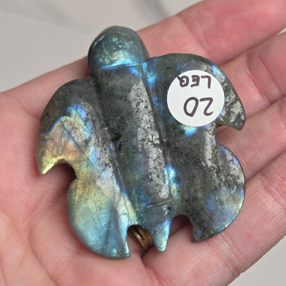 A hand holding a polished labradorite stone charm shaped like a stylized animal head, showing iridescent blue, green, and gold flashes on a dark gray base. A small white sticker with the handwritten text '20 LEG' is attached to the piece.
