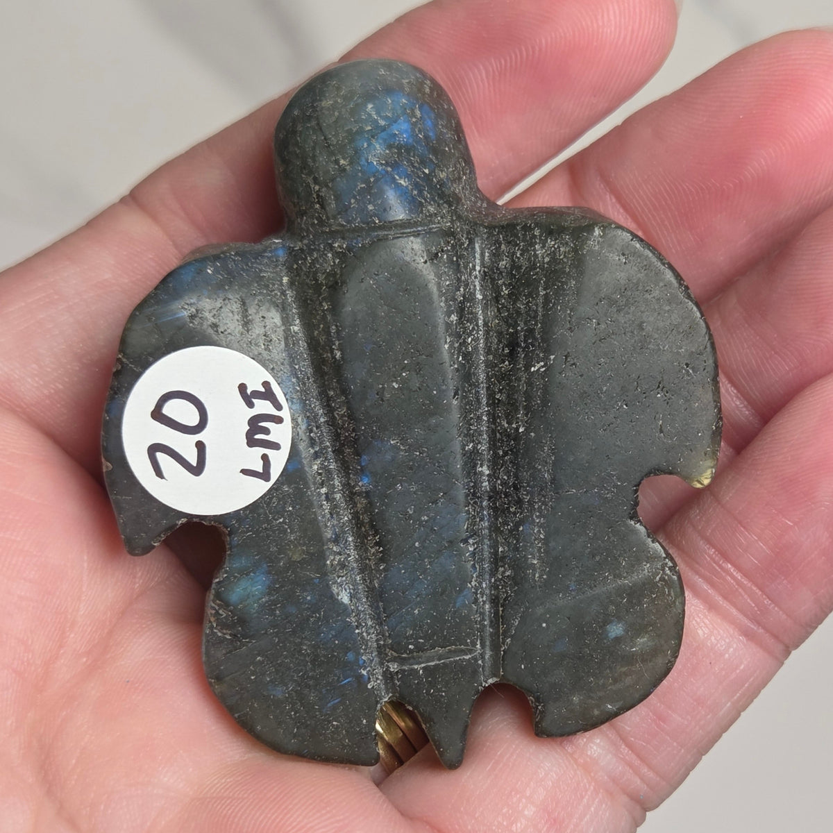 A hand holding a small carved stone figurine with a white sticker reading '20 LME' on it, photographed against a light neutral background.