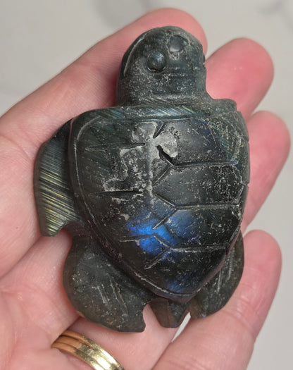 A hand holding a carved sea turtle figurine made of dark stone with a blue iridescent sheen on its shell, set against a light marble background.