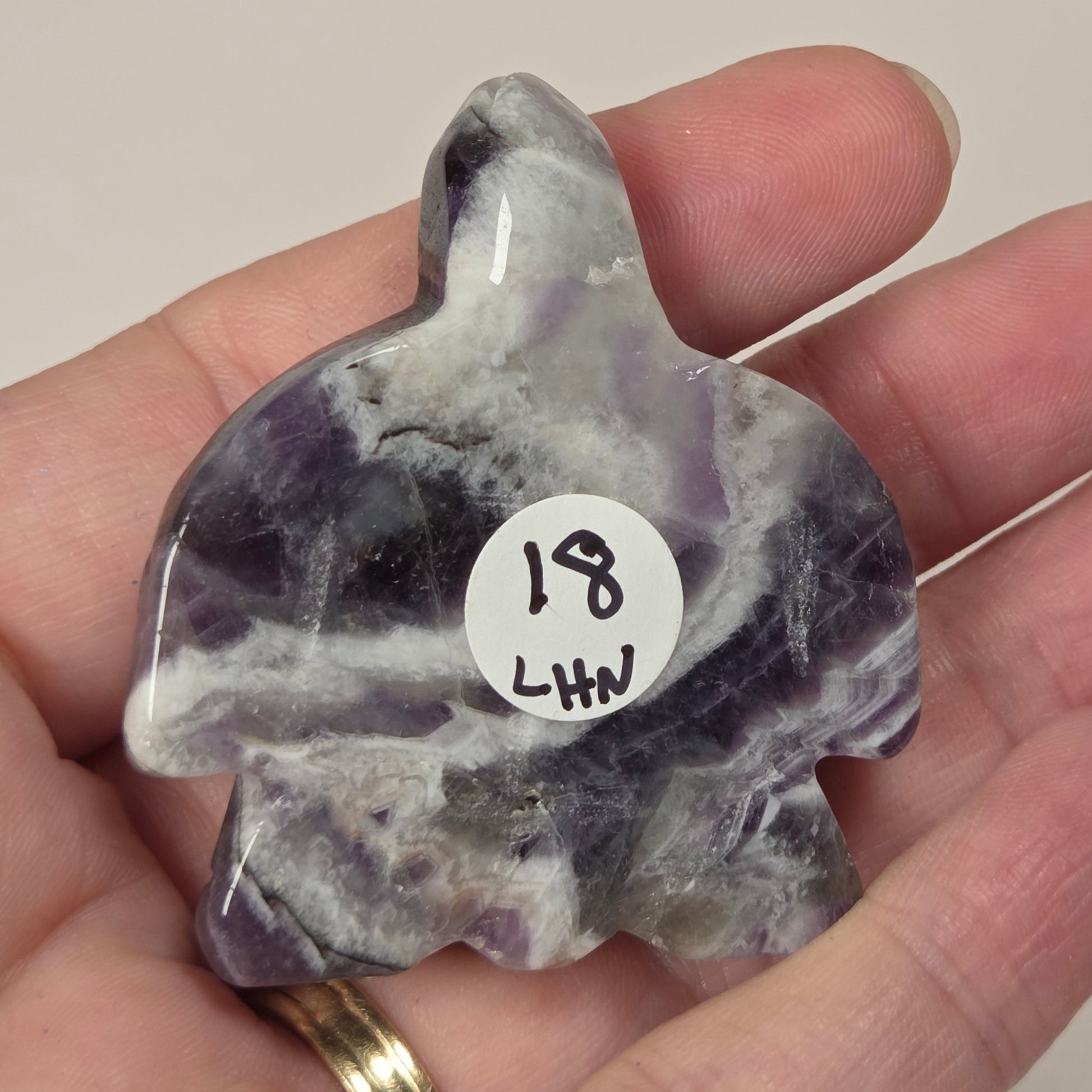 A hand holds a polished amethyst turtle-shaped stone with a white circular sticker marked '18 LHW'. The stone features natural purple and white crystalline patterns.
