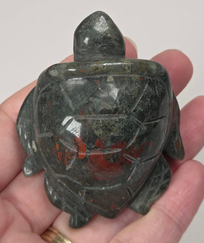 A carved turtle figurine made of dark green stone with red matrix inclusions, held in a person's hand against a plain light background.