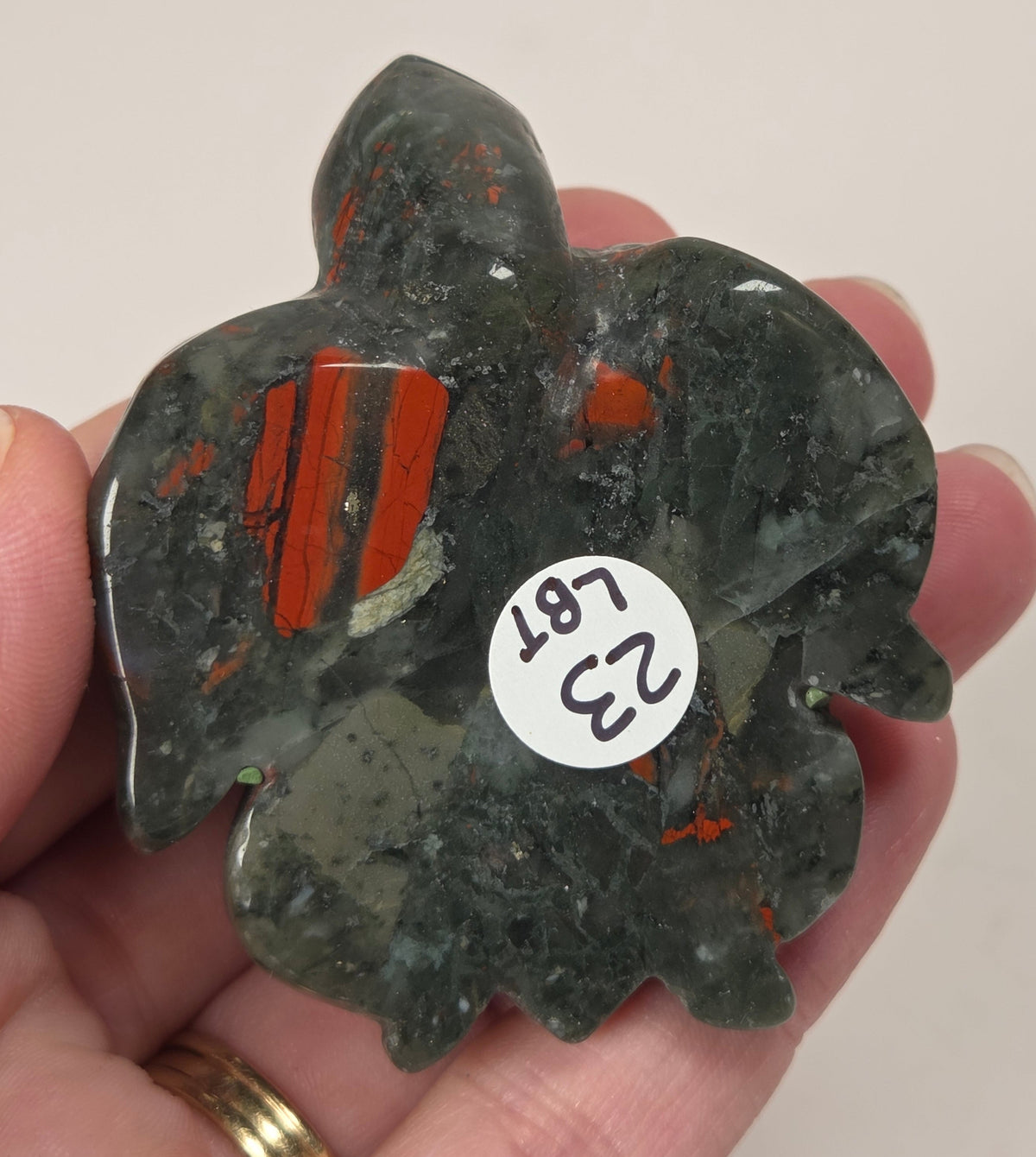A hand holds a carved dark green stone pendant with orange-red inclusions and a small white sticker labeled '23 LB 171'.