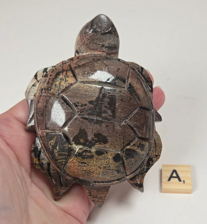 A hand holds a small decorative turtle figurine with a glossy brown and black shell pattern, shown next to a small wooden letter tile on a plain white background.