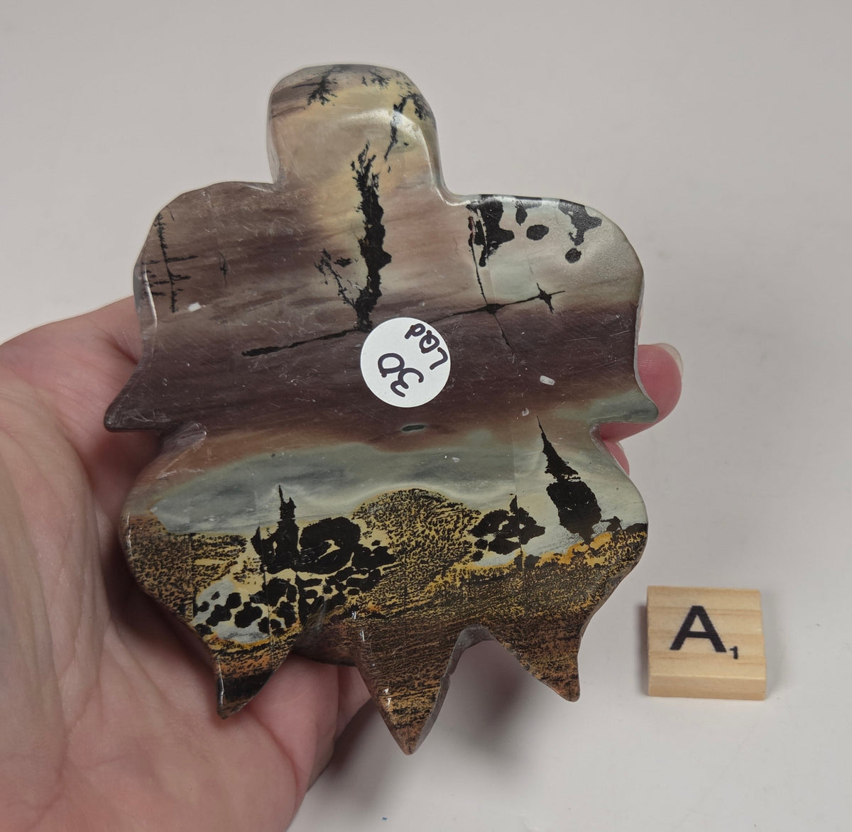 A hand holds a decorative, hand-carved wooden plaque shaped like a stylized figure, featuring a detailed landscape scene with black trees and birds on a brown and beige background. A small wooden letter tile with the letter 'A' sits next to it on a plain light gray surface.
