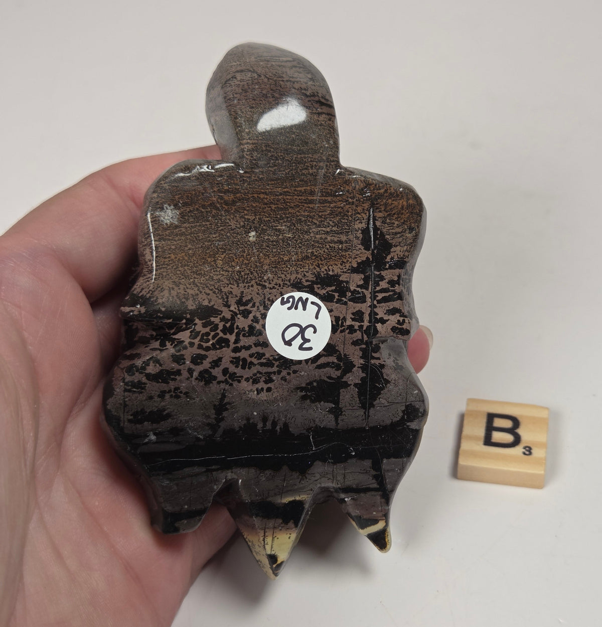 A hand holds a small, rustic, hand-carved wooden object with a ghost-like silhouette, featuring a glossy dark brown and black textured finish. A small white inventory sticker is attached to the side, and a wooden Scrabble tile spelling 'B's' sits nearby on a plain off-white background.