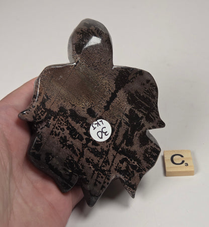 A hand holds a dark, irregularly shaped ceramic piece with a glossy brown and black marbled glaze. A small wooden tile with the letter 'C' sits nearby on a plain white surface.